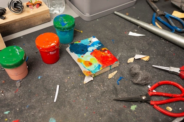 A workbench cluttered with various cosplay materials, including EVA foam sheets, PVC pipes, acrylic paints in vibrant colors, an assortment of glass and plastic vials, leather straps, buckles, and crafting tools like scissors, glue guns, and sandpaper.